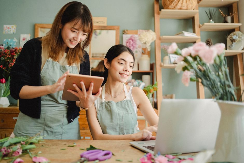 Two women working in a florist shop, with one standing and holding a tablet while smiling, and the other seated at a wooden worktable with a silver laptop in front of her. The seated woman has dark ha
