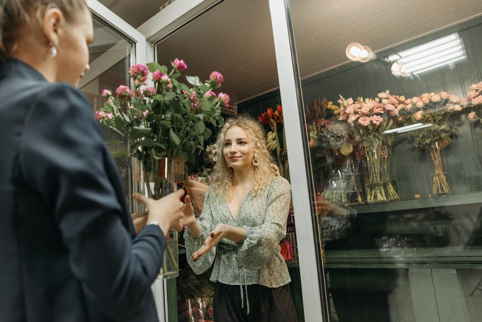 A young woman with curly blonde hair and light skin stands inside a flower shop, engaging in a floral transaction. She is wearing a light green, floral-patterned blouse with long sleeves and black tro