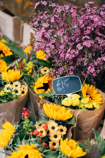 A vibrant display of fresh floral arrangements at a flower market, featuring a predominance of bright yellow sunflowers and smaller daisies, wrapped in brown paper and green foliage. Behind the sunflo