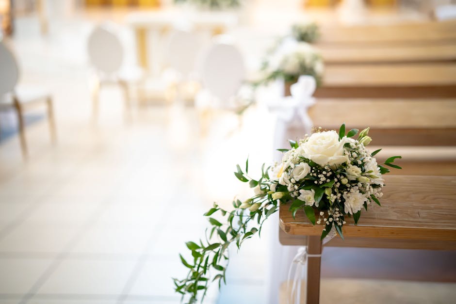 A white floral arrangement consisting of roses, baby's breath, and other small white blooms with green foliage, placed on the edge of a wooden pew in a bright, softly lit interior setting. The bouquet