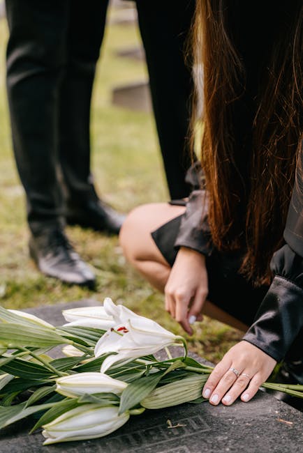 A floral tribute featuring white lilies with large, open petals and prominent red stamens, arranged with green leaves and stems. The flowers are laid on a dark stone memorial, and a woman with long, b