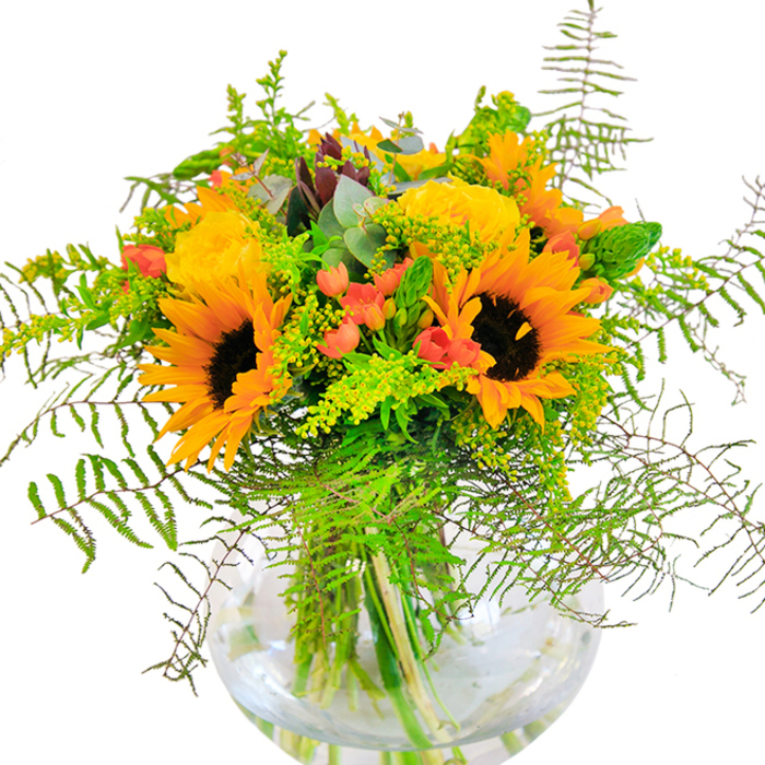 At first glance, this bouquet exudes sunlight, joy, and the lively character that defines Chadwell Heath's close-knit community. Luminous sunflowers with inky brown centers emerge at the heart of the design, their golden petals unfurling like rays over the tables of local homes. Intertwined with them, tightly packed yellow roses spiral open, their elegant texture reminiscent of classic arrangements gracing family celebrations around Ashton Gardens. Sprays of glistening scarlet hypericum berries inject lively contrast, sparkling among layers of fresh, deep green foliage. The cascading ferns and airy filler greens capture the easy grace of nearby green spaces, while subtle blue-green eucalyptus leaves lend a whisper of cool, modern freshness. The clear, round glass vase perfectly cradles the bouquet's vibrant stems, their lines magnified in the shimmering water-highlighting the structural harmony prized by discerning florists. One can almost imagine the delicate scent that hovers above the arrangement: a blend of sweet rose, earthy sunflower, and the crispness of the outdoors. This composition, alive with texture and colour, is ideal for elevating landmark occasions-a joyful birthday, anniversary, or simply welcoming loved ones home. Crafted by the skilled artisans at Florist Chadwell Heath, this bouquet is a radiant centerpiece that infuses any moment with happiness and the unmistakable warmth of the local community.